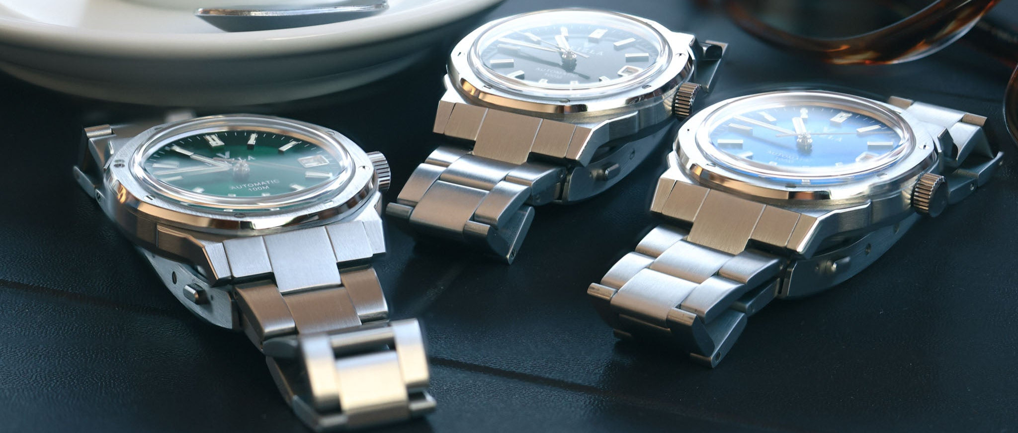 Three silver watches on a dark surface with a cup and spoon in the background.